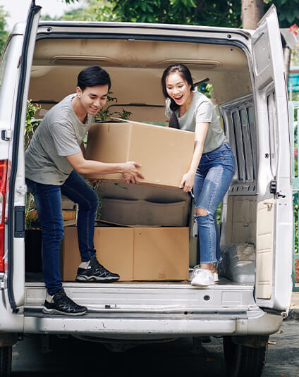 A smiling young man and woman are lifting a large cardboard box into the back of a white moving van, with several other boxes already stacked inside the vehicle.