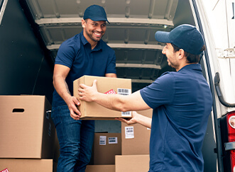Two smiling couriers in blue uniforms and caps are unloading cardboard packages from the back of a delivery van. One courier stands inside the van handing a box to the other courier standing outside.