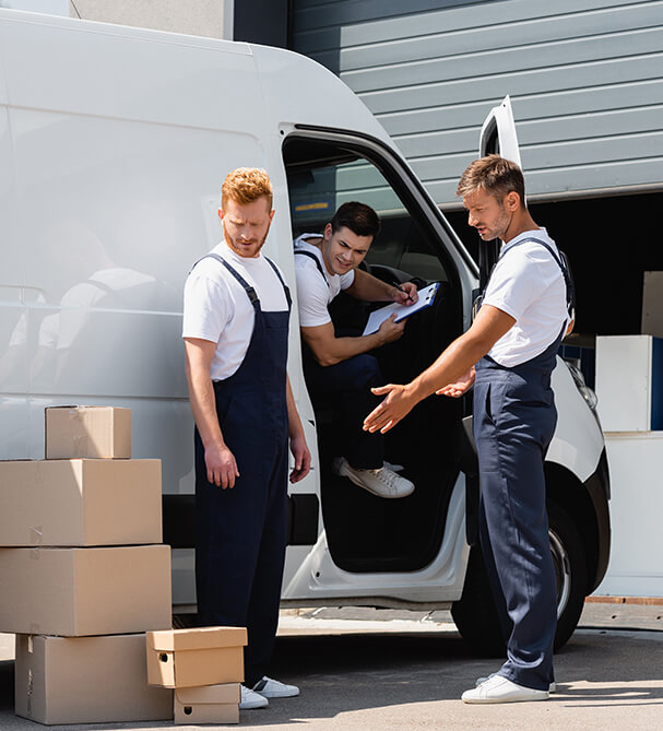 Three movers in blue overalls and white t-shirts work around a white delivery van. One stands beside a stack of cardboard boxes, looking down; another sits inside the van, writing on a clipboard; and the third stands outside the van, gesturing toward the boxes.