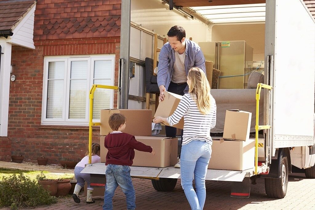 A family consisting of a man, woman, young boy, and girl unloads cardboard boxes from the back of a moving truck parked in front of a brick house. The man passes a box to the woman, while the children stand near other boxes on the truck’s ramp, assisting with the move.
