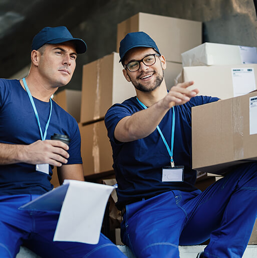 Two male delivery workers in blue uniforms and caps stand inside a truck filled with cardboard boxes. One holds a box and points, while the other holds a coffee cup and looks ahead.