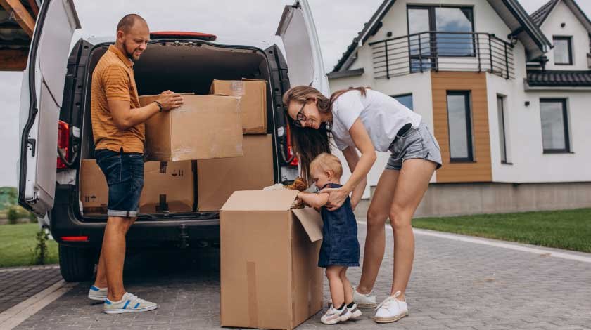 A family, including a man, woman, and toddler, is unloading boxes from a white moving van in front of a modern house. The man lifts a large cardboard box from the van, while the woman and toddler stand near a box on the ground, with the toddler appearing to engage with it, suggesting a moving day scene.