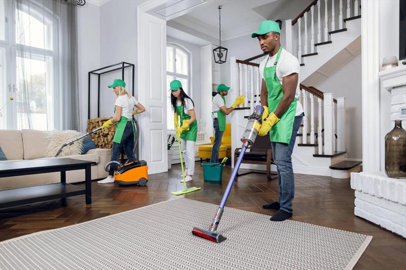 A team of professional cleaners in green uniforms and yellow gloves clean a modern living room—one vacuums a rug, another a sofa, while two others mop and clean surfaces.