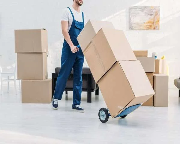A person in blue overalls uses a hand truck to move stacked cardboard boxes across a light-colored floor, with more boxes and furniture in the room.