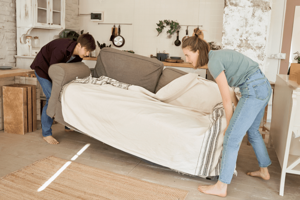 A man and woman carry a large light-colored sofa with a patterned throw, moving it through a home with a kitchen visible in the background.