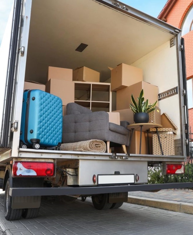 The back of a moving truck loaded with boxes, furniture, a blue suitcase, and a plant, ready for a house move.