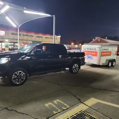 A black Toyota Tundra hitched to a white U-Haul trailer is parked at a U-Haul facility at night, with illuminated signage and asphalt ground.