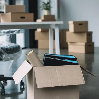 A partially packed office with a plastic-wrapped chair in the foreground and boxes stacked on a desk and floor, suggesting a relocation.