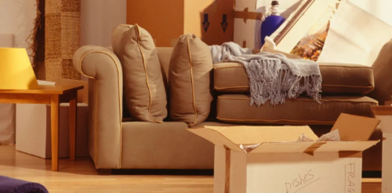 A living room with moving boxes, a couch, and a table with a lamp, including a labeled box marked "Dishes," suggesting residential moving.