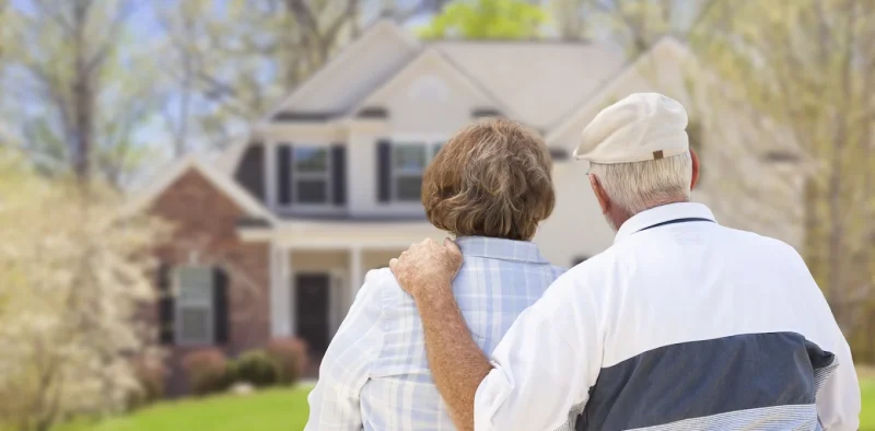 An elderly couple sits arm-in-arm outdoors, viewed from behind, facing a blurred two-story house in the background.