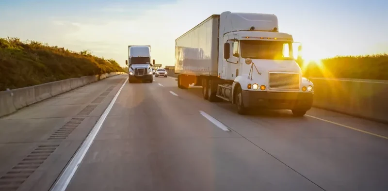 A white semi-truck drives on a multi-lane highway at sunset, with another truck in the distance and a grassy embankment on the left.