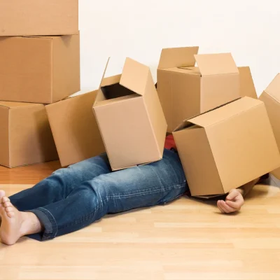 A person lies overwhelmed on a light wooden floor, buried under cardboard boxes, with more stacked in the background.