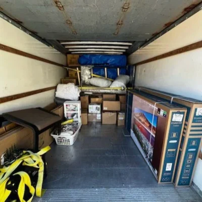 The interior of a moving truck packed with boxes, furniture, TVs in original packaging, and moving straps, indicating a relocation.