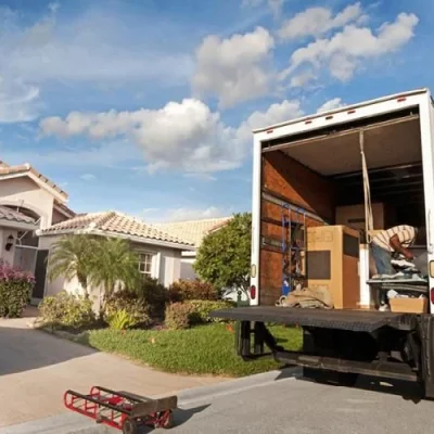 A moving truck with its ramp down is parked by a house, showing boxes and furniture inside. A hand truck lies on the driveway near a parked car.