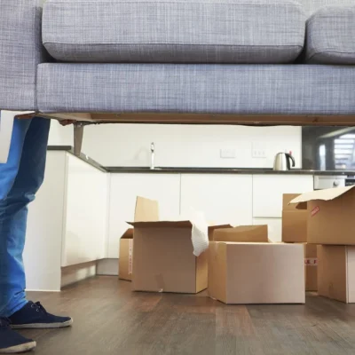 A person in jeans and a plaid shirt carries a gray sofa across a wooden floor in a room with moving boxes and a visible kitchen.