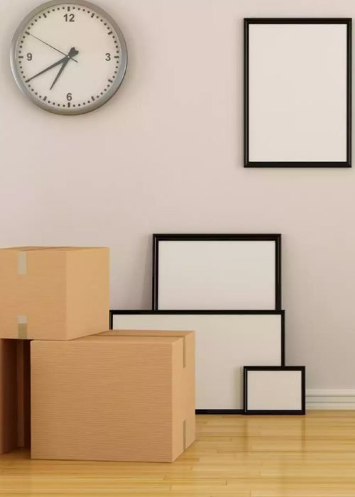 A room being prepared for moving, with a stack of cardboard moving boxes, several empty picture frames leaning against a wall, a wall clock, a white radiator, and a green ladder positioned nearby. The space has light-colored walls and wooden flooring.