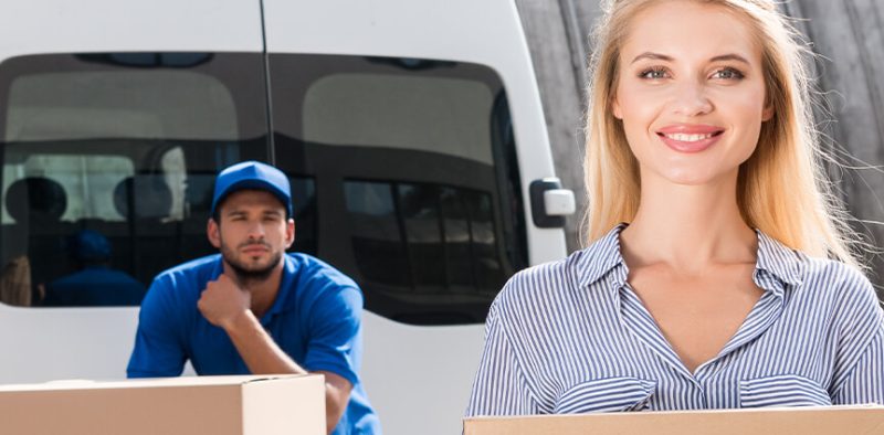 A smiling woman holds a package in the foreground, suggesting she has just received a delivery. In the background, a delivery driver in a blue uniform sits in the back of a white delivery van, also looking towards the camera. The scene conveys a successful and friendly package delivery.
