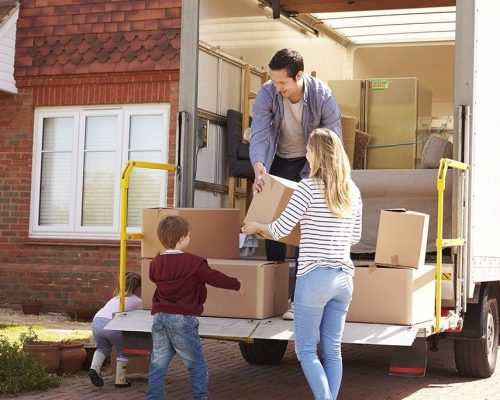 A family consisting of a man, woman, young boy, and girl unloads cardboard boxes from the back of a moving truck parked in front of a brick house. The man passes a box to the woman, while the children stand near other boxes on the truck’s ramp, assisting with the move.