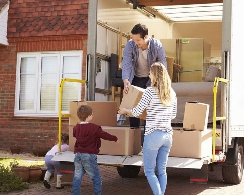 A family consisting of a man, woman, young boy, and girl unloads cardboard boxes from the back of a moving truck parked in front of a brick house. The man passes a box to the woman, while the children stand near other boxes on the truck’s ramp, assisting with the move.