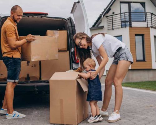 A family, including a man, woman, and toddler, is unloading boxes from a white moving van in front of a modern house. The man lifts a large cardboard box from the van, while the woman and toddler stand near a box on the ground, with the toddler appearing to engage with it, suggesting a moving day scene.