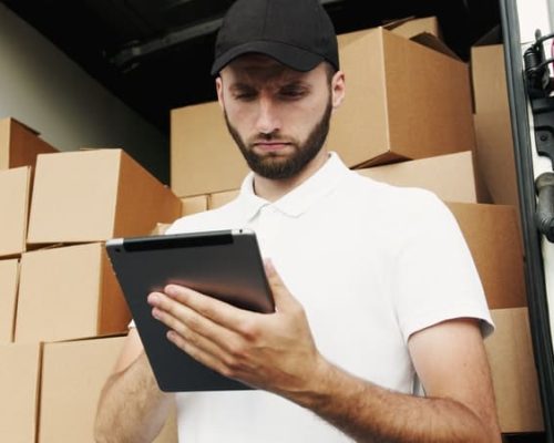 A delivery driver wearing a white polo shirt and black cap stands inside the back of a delivery truck filled with cardboard boxes, looking down at a tablet in his hands.