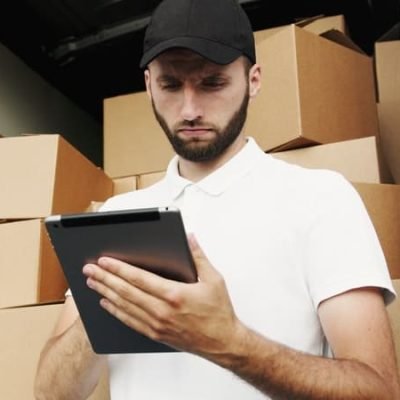A delivery driver wearing a white polo shirt and black cap stands inside the back of a delivery truck filled with cardboard boxes, looking down at a tablet in his hands.