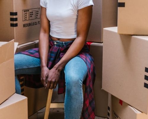 A person sitting on a stool in a room surrounded by moving boxes, including one marked with a red "FRAGILE" label, indicating an active packing or unpacking process during a move.