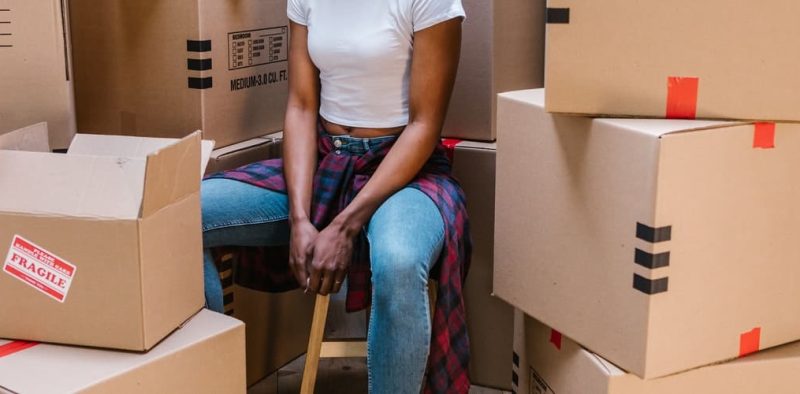 A person sitting on a stool in a room surrounded by moving boxes, including one marked with a red "FRAGILE" label, indicating an active packing or unpacking process during a move.