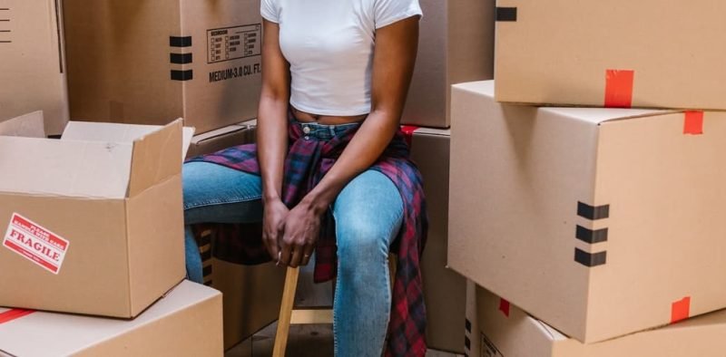 A person sitting on a stool in a room surrounded by moving boxes, including one marked with a red "FRAGILE" label, indicating an active packing or unpacking process during a move.